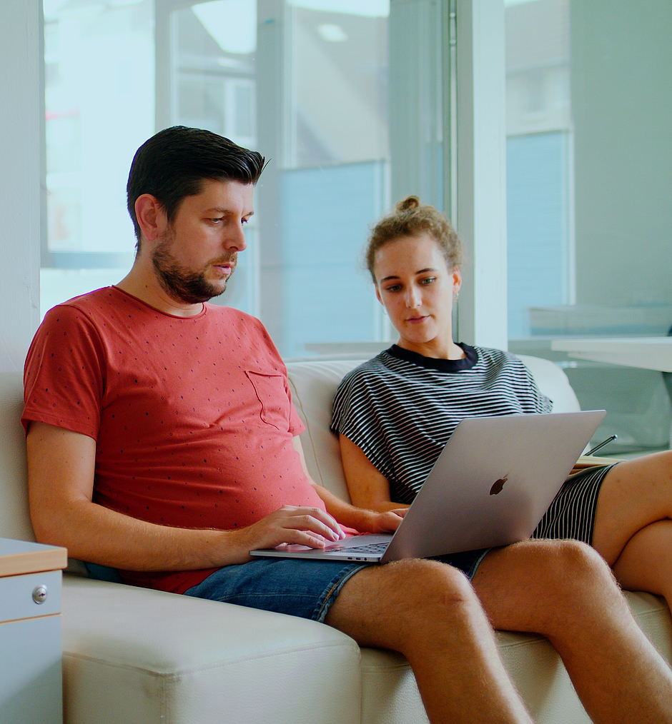 Zwei Mitarbeitende sitzen auf einem hellen Sofa mit einem Laptop